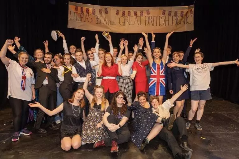 The cast of utopia are assembled in several lines looking towards the camera. A banner reading "Utopia's 35th Annual Great British Picnic" in large text hangs above them.